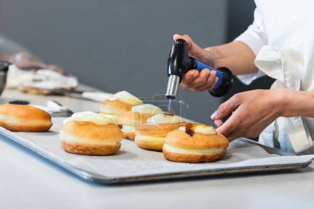 Foto de Chef de cultivo irreconocible caramelizante crema de azúcar en berlineses dulces con antorcha de gas mientras prepara postre vegano en panadería - Imagen libre de derechos