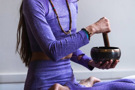 Téléchargez les photos : Cultivé femme méconnaissable en vêtements de sport assis dans la pose Lotus et jouer bol de chant tibétain tout en méditant et en pratiquant le yoga - en image libre de droit