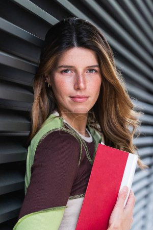 Téléchargez les photos : Jeune femme de contenu dans des vêtements décontractés tenant le manuel tout en s'appuyant contre le mur nervuré en ville pendant la journée - en image libre de droit