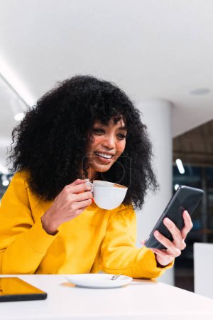 Foto de Contenido joven étnica femenina con el pelo largo Afro sonriendo y navegando por el teléfono móvil mientras bebe taza de café en la cafetería ligera - Imagen libre de derechos