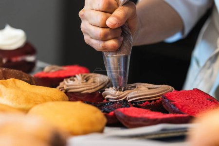 Téléchargez les photos : Crop anonyme cuisinier avec sac à pâtisserie en ajoutant de la crème au chocolat sur les gâteaux éponges végétaliens tout en préparant des desserts dans la boulangerie - en image libre de droit