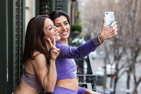 Photo for Side view of happy young Hispanic woman smiling with friend making piece gesture while taking selfie on smartphone standing on balcony - Royalty Free Image