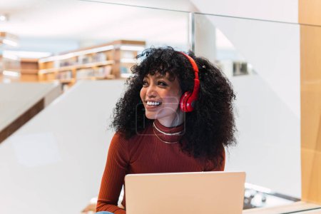 Téléchargez les photos : Femme aux cheveux bouclés noirs avec écouteurs regardant loin avec sourire tout en étant assis avec un ordinateur portable dans la bibliothèque moderne - en image libre de droit
