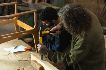 Foto de Alto ángulo de carpinteros especializados concentrados con martillo y destornillador que hacen silla mientras trabajan en carpintería - Imagen libre de derechos