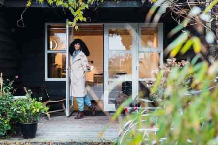 Foto de Joven mujer afroamericana alegre con el pelo oscuro rizado en un abrigo caliente sosteniendo una taza de café y saliendo de la casa a través de la puerta de cristal y mirando a la cámara - Imagen libre de derechos