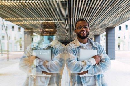 Photo for Smiling African American male with modern netbook in hands looking at camera while standing near glass wall of modern building on street - Royalty Free Image