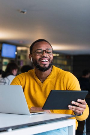 Foto de Hombre negro feliz sentado en la mesa con portátil y escuchando música en los auriculares de la tableta en el espacio de coworking - Imagen libre de derechos