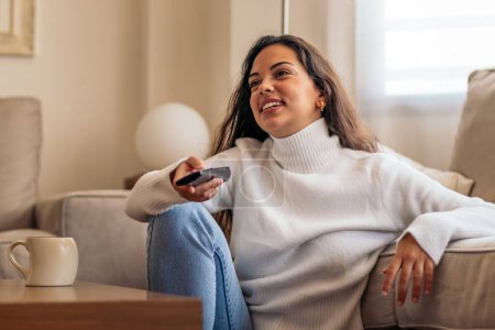 mujer joven sentada en su sofá de la casa, sosteniendo el control remoto de la televisión mientras disfruta de un programa. está disfrutando de su tiempo libre en casa