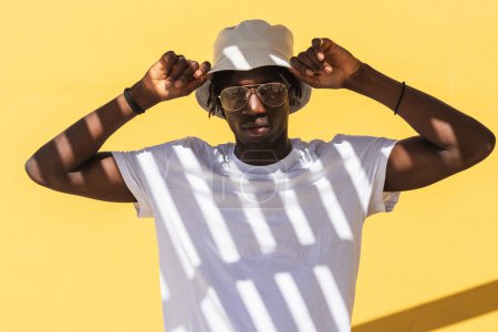 Photo for Modern hipster African American guy with striped shadow on white t shirt wearing sunglasses and panama hat and looking at camera while standing against yellow wall - Royalty Free Image