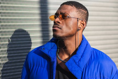 Photo for Confident African American male wearing trendy orange sunglasses and warm jacket standing near metal wall of modern building in city and looking away - Royalty Free Image