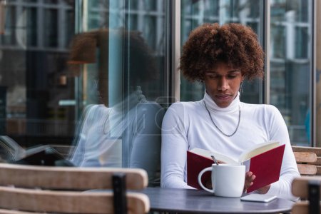 Téléchargez les photos : Calme jeune Afro-Américain aux cheveux bouclés en tenue tendance reposant sur la terrasse du café avec une tasse de café et la lecture livre intéressant - en image libre de droit