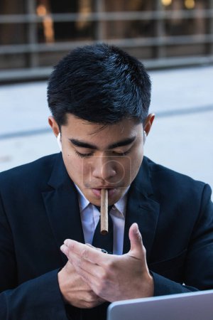 Foto de Confident Asian male entrepreneur in formal clothes lighting up cigar in city street - Imagen libre de derechos