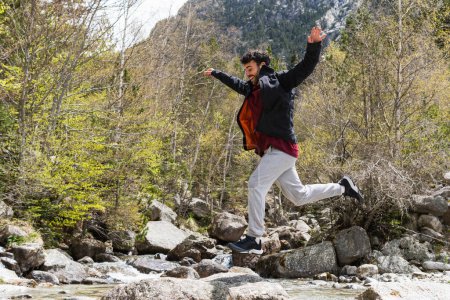 Téléchargez les photos : Vue latérale du voyageur masculin ravi sautant par-dessus le ruisseau pendant les vacances dans les Pyrénées - en image libre de droit