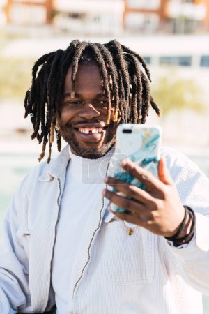 Stylish African American male with dreadlocks and in trendy outfit taking self portrait on smartphone in city on sunny day