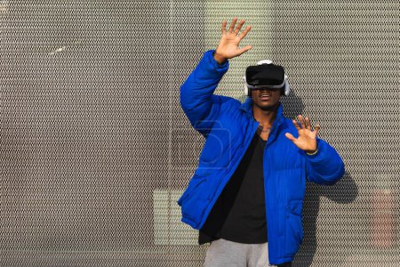 Téléchargez les photos : Homme afro-américain debout sur la rue près d'une clôture métallique tout en explorant la réalité virtuelle dans des lunettes innovantes et toucher l'air - en image libre de droit