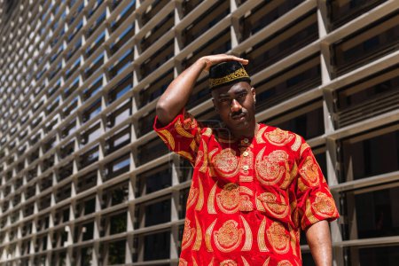 Ernsthafter erwachsener afrikanischer Typ in traditioneller roter Kleidung und Kufi-Mütze, der in die Kamera blickt, während er an sonnigen Tagen in der Stadtstraße in der Nähe eines Gebäudes mit Glaswänden steht
