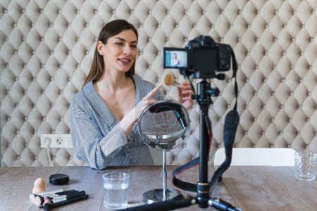 Cheerful attractive brunette in casual dress applying powder with brush on face while recording vlog video on camera