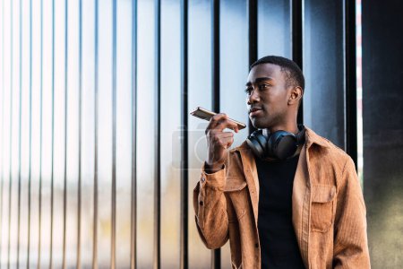 Téléchargez les photos : Homme afro-américain sérieux avec écouteurs sans fil noirs enregistrant un message vocal sur un smartphone tout en se tenant près d'un mur métallique en ville - en image libre de droit