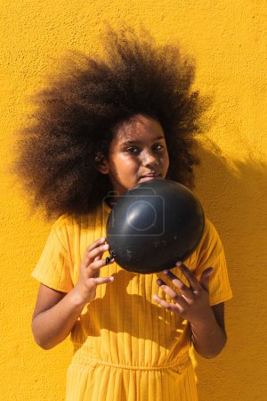 Téléchargez les photos : Afro-américaine adolescent fille avec bouclé coiffure tenant ballon noir tout en se tenant sur fond jaune regardant loin - en image libre de droit