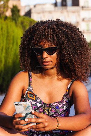 Photo for Young black woman in trendy outfit and sunglasses sitting cross legged on border and browsing smartphone on summer day - Royalty Free Image