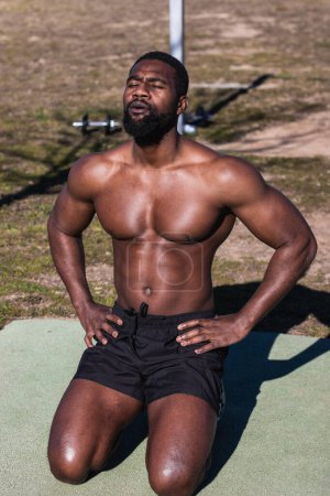 Photo for Shirtless muscular black male athlete with closed eyes breathing while sitting on knees with hands on hips during workout in sunlight - Royalty Free Image