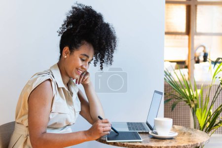 Foto de Vista lateral de una mujer afroamericana freelancer positiva usando tableta gráfica en la mesa con computadora portátil y café - Imagen libre de derechos