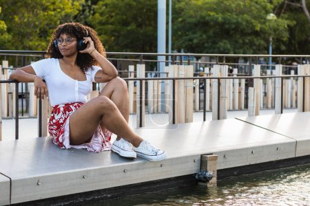 Photo for Side view of stylish African American female listening to music in headphones while resting near water in city - Royalty Free Image