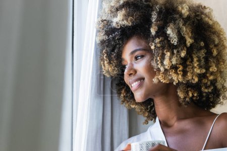 Foto de Pacifica mujer afroamericana encantada con el pelo rizado con camisa blanca de pie cerca de la ventana con taza de bebida en la mañana - Imagen libre de derechos