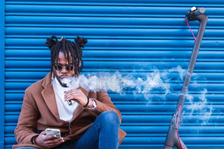 Photo for Modern adult hipster African American male in trendy outfit smoking electronic cigarette and browsing mobile phone while sitting near electric scooter against blue wall - Royalty Free Image