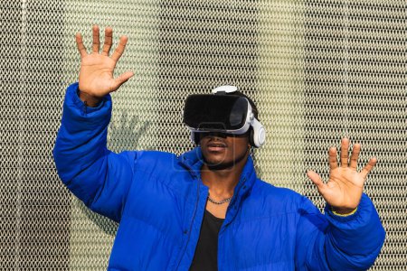 Afroamerikaner steht auf Straße in der Nähe von Metallzaun und erkundet virtuelle Realität mit innovativer Brille und Berührung der Luft