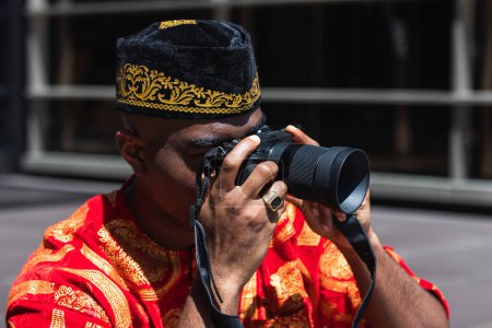 Anonymous black man in traditional red clothes and kufi taking picture on professional photo camera in sunny day in city street