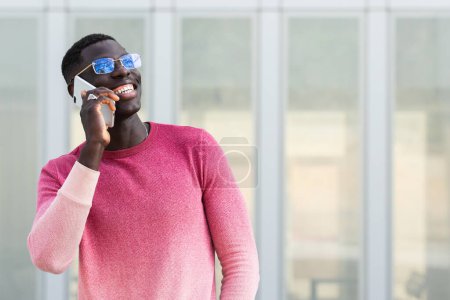 Foto de Joven empresario afroamericano en gafas que habla por celular mientras mira a la luz del día - Imagen libre de derechos