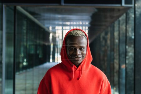 Téléchargez les photos : Jeune homme afro-américain millénial avec capuche ajustable teint et regardant la caméra dans le passage moderne - en image libre de droit