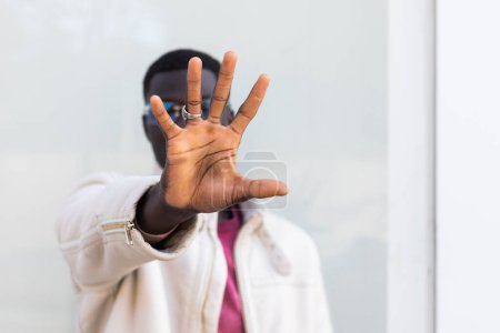 Téléchargez les photos : Homme afro-américain en vêtements à la mode et lunettes de soleil face cachée et tendre la main vers la caméra tout en montrant le panneau stop contre le bâtiment urbain - en image libre de droit