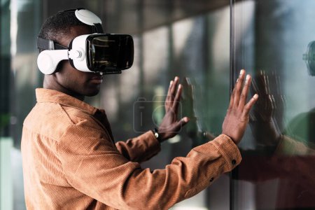 Positiver afroamerikanischer Mann mit moderner VR-Brille steht neben Glaswand und erkundet virtuelle Realität auf der Straße der Stadt