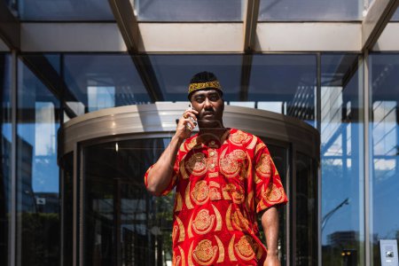 Ernsthafter erwachsener schwarzer Mann in traditioneller roter Kleidung und Kufi steht in der Nähe eines Gebäudes mit Glaswänden, während er an sonnigen Tagen in der Stadtstraße telefoniert und wegschaut