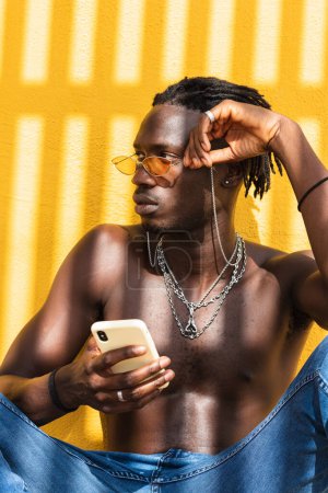 Photo for Serious young shirtless African American man in trendy sunglasses and with metal chains browsing mobile phone while sitting against yellow wall with striped shadow - Royalty Free Image