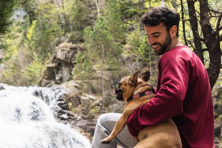 Delighted male hiker embracing French Bulldog near waterfall in woods while enjoying trekking in Pyrenees
