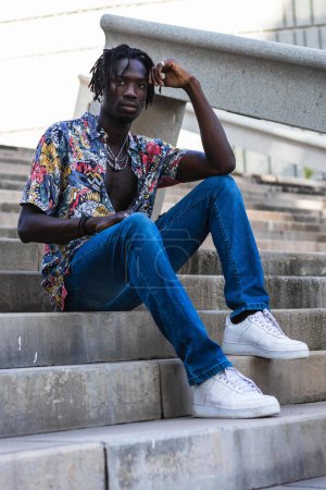 Photo for Low angle full body of serious African American male in colorful shirt and jeans with sneakers sitting on stone steps and looking at camera on urban street - Royalty Free Image
