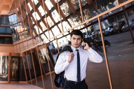 Téléchargez les photos : Entrepreneur masculin joyeux en costume formel noir debout sur les escaliers près du bâtiment moderne et discuter des questions d'affaires sur le téléphone mobile tout en regardant loin - en image libre de droit