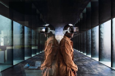 Photo for Side view of African American male in modern VR goggles standing backwards a glass wall with reflection while exploring virtual reality - Royalty Free Image