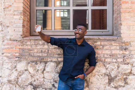 Foto de Feliz hombre adulto negro en gafas y camisa tomando selfie en el teléfono celular en la calle de la ciudad en la luz del día cerca de edificio de ladrillo con ventana - Imagen libre de derechos