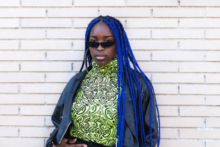 Selbstbewusste Afroamerikanerin mit blauen Zöpfen im schicken Outfit und Sonnenbrille steht auf der Straße und blickt in die Kamera