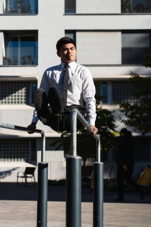 Photo for Asian male entrepreneur in formal outfit balancing on parallel bars while doing abs exercises and grimacing - Royalty Free Image