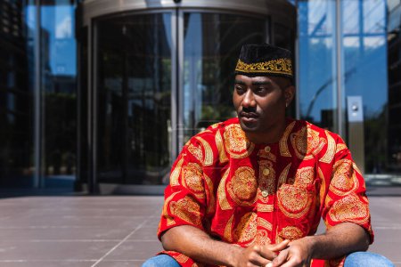 Ernsthafter erwachsener afrikanischer Typ in traditioneller roter Kleidung und Kufi-Mütze, der bei sonnigem Wetter in der Stadtstraße in der Nähe eines Gebäudes mit Glaswänden sitzt und wegschaut
