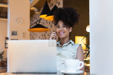 Photo for Positive African American female with Afro hairstyle talking on mobile phone at table with netbook and cup of coffee - Royalty Free Image