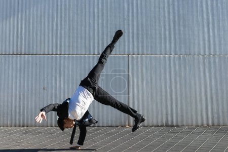 Téléchargez les photos : Asiatique entrepreneur masculin en costume formel montrant mouvement de breakdance et équilibrage sur le bras dans le handstand sur la rue de la ville tout en regardant la caméra - en image libre de droit