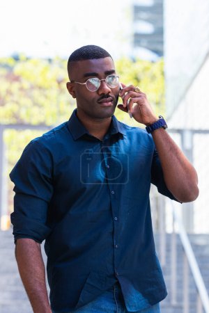 Photo for Confident African American adult guy in shirt and eyeglasses standing in city street while having conversation on mobile phone near stairs with railings in daylight - Royalty Free Image