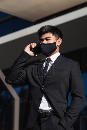 Foto de Vista lateral de hombre de negocios asiático en máscara protectora y traje elegante de pie en la ciudad y tener conversación de teléfono inteligente mientras mira hacia otro lado - Imagen libre de derechos