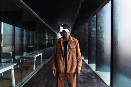 Hombre afroamericano en gafas VR explorando el ciberespacio mientras está parado en la calle cerca de un edificio moderno con pared de vidrio